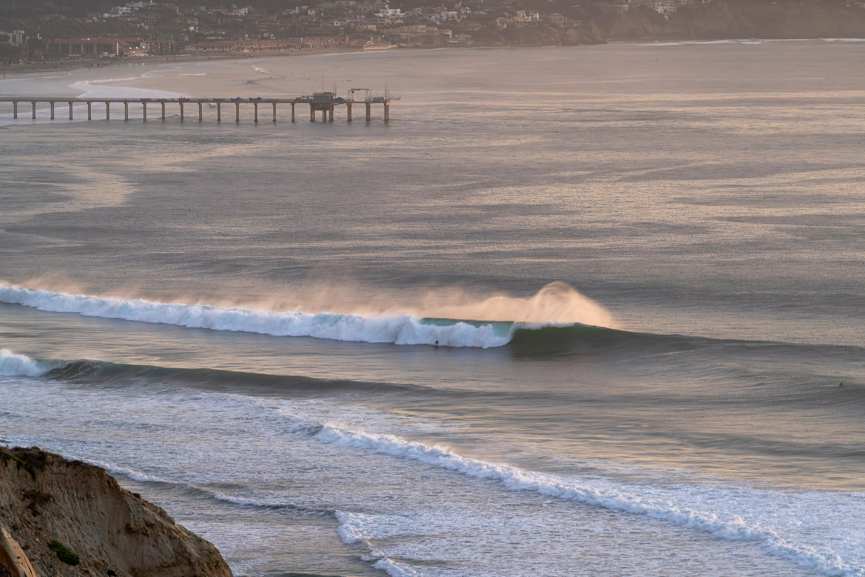Waves crashing against a beach with a pier in the distance.
