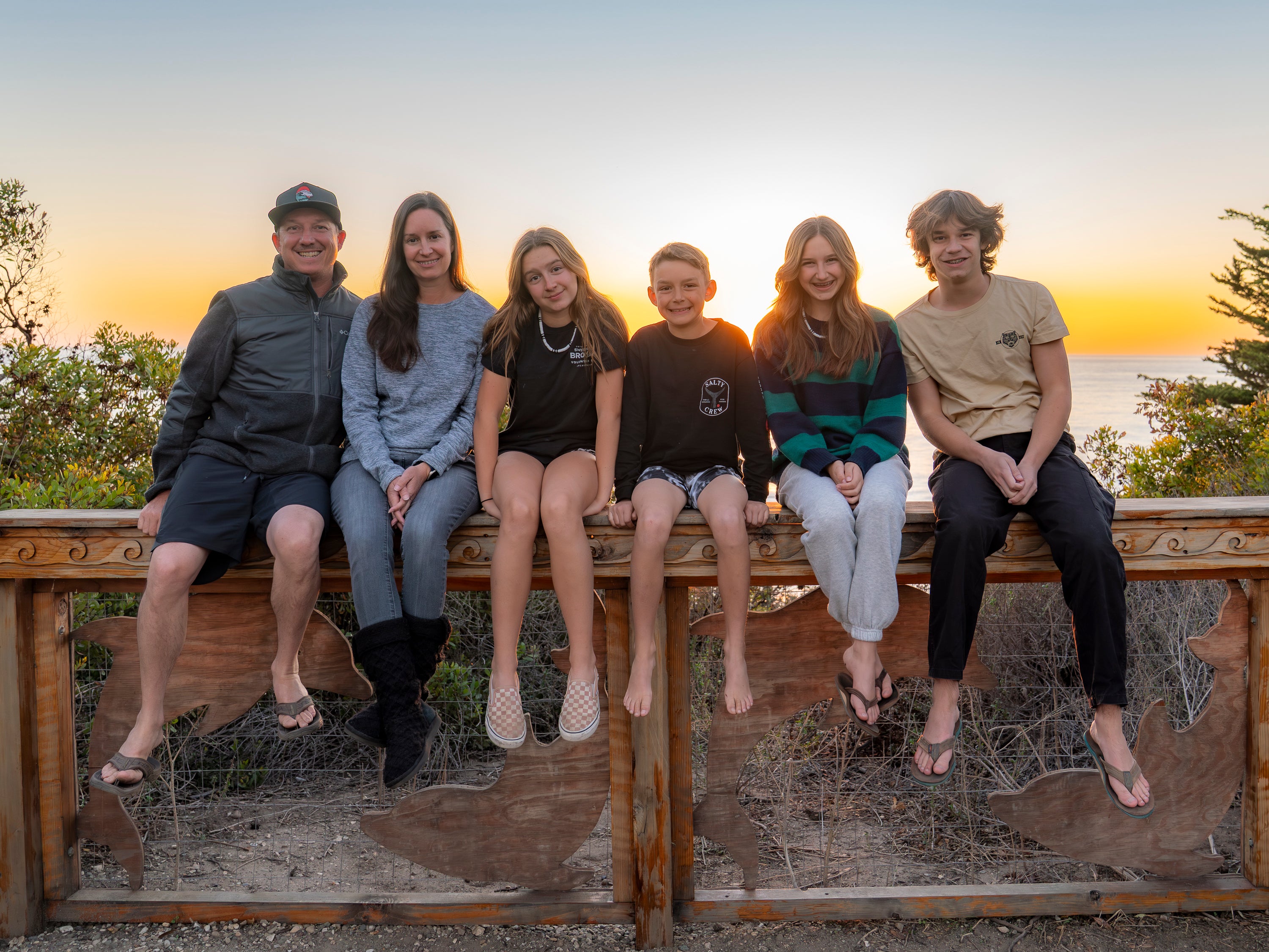 Family of six sitting on a wooden railing with a scenic background. El Capitan Beach, Santa Barbara, California.