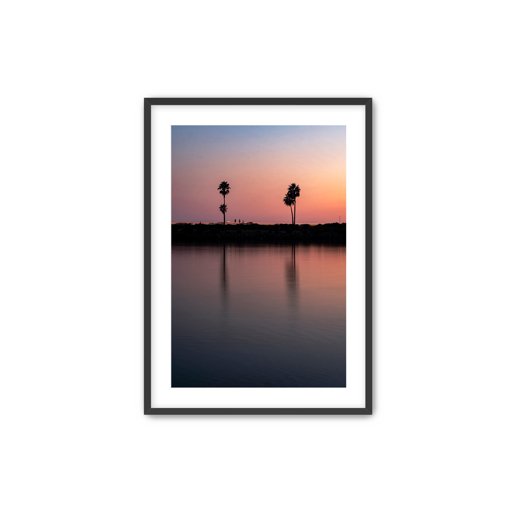 Framed artwork of a sunset over a Carlsbad lagoon with palm trees and cars on highway 101 in the background, placed on a white wall over a desk.