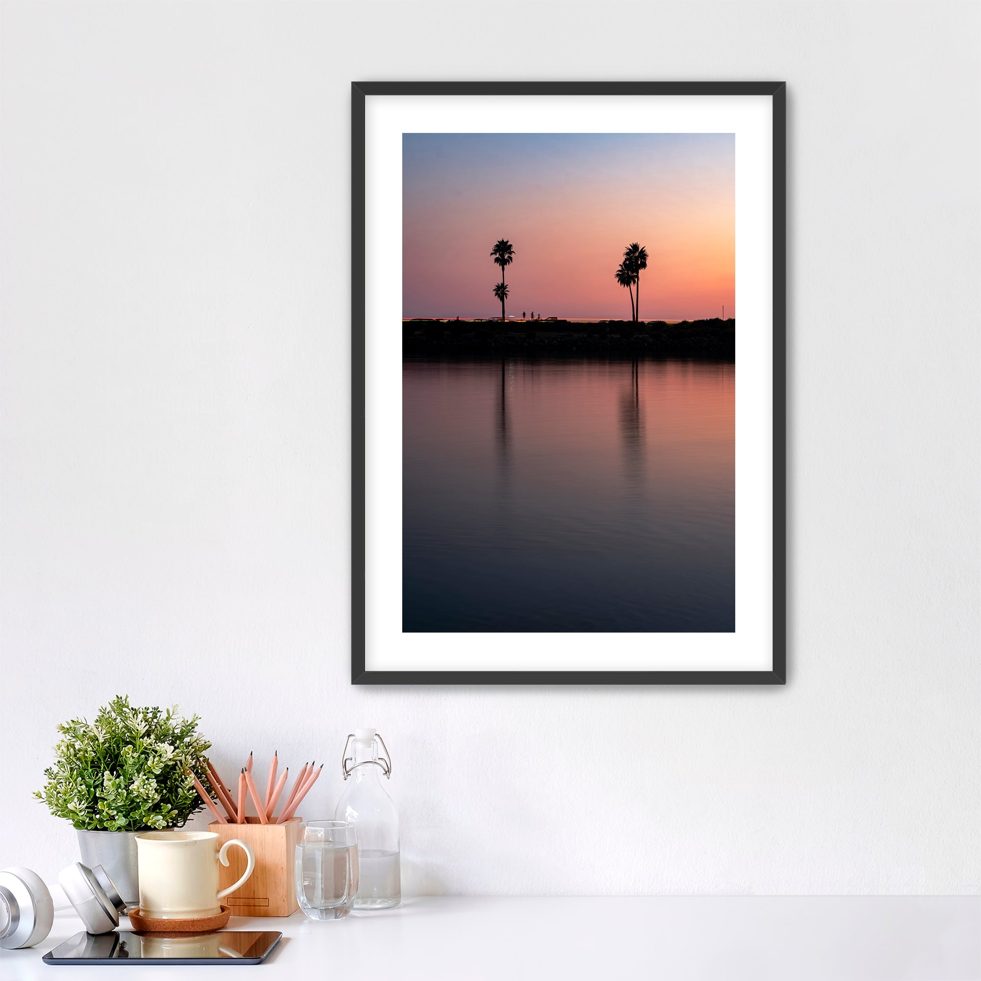 Framed artwork of a sunset over a Carlsbad lagoon with palm trees and cars on highway 101 in the background, placed on a white wall over a desk.