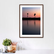 Framed artwork of a sunset over a Carlsbad lagoon with palm trees and cars on highway 101 in the background, placed on a white wall over a desk.