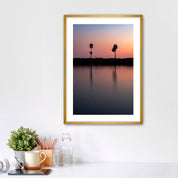 Framed artwork of a sunset over a Carlsbad lagoon with palm trees and cars on highway 101 in the background, placed on a white wall over a desk.