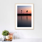 Framed artwork of a sunset over a Carlsbad lagoon with palm trees and cars on highway 101 in the background, placed on a white wall over a desk.