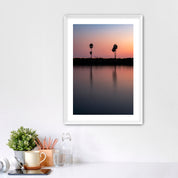Framed artwork of a sunset over a Carlsbad lagoon with palm trees and cars on highway 101 in the background, placed on a white wall over a desk.