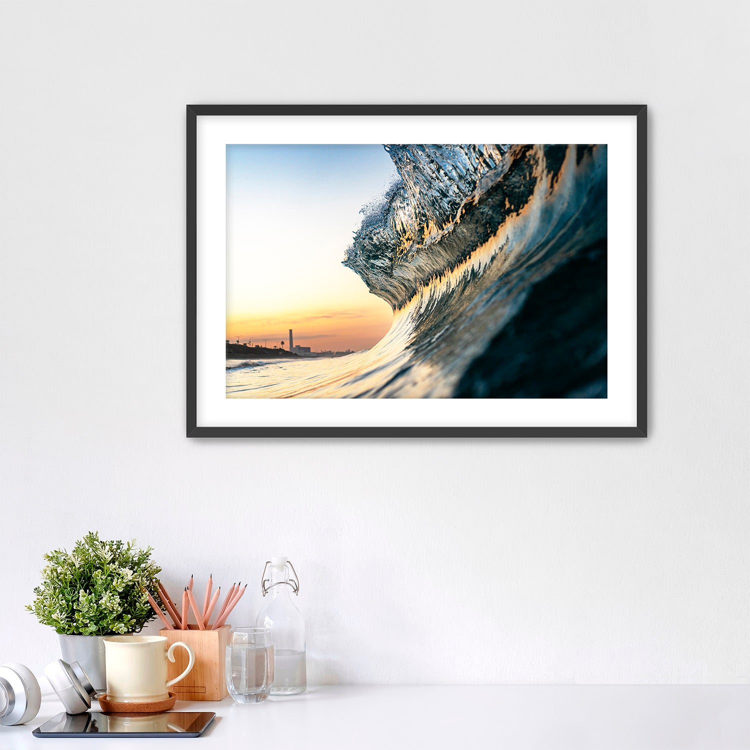 Framed photograph of a wave crashing on a beach with a power plant in the distance, displayed on a wall above a desk.
