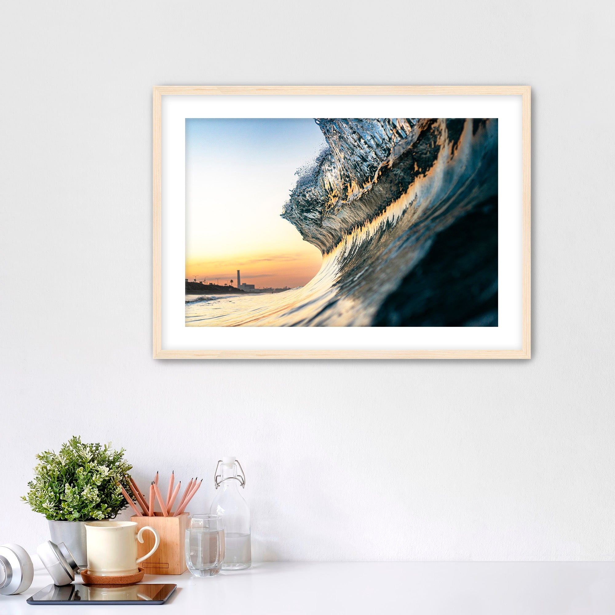 Framed photograph of a wave crashing on a beach with a power plant in the distance, displayed on a wall above a desk.