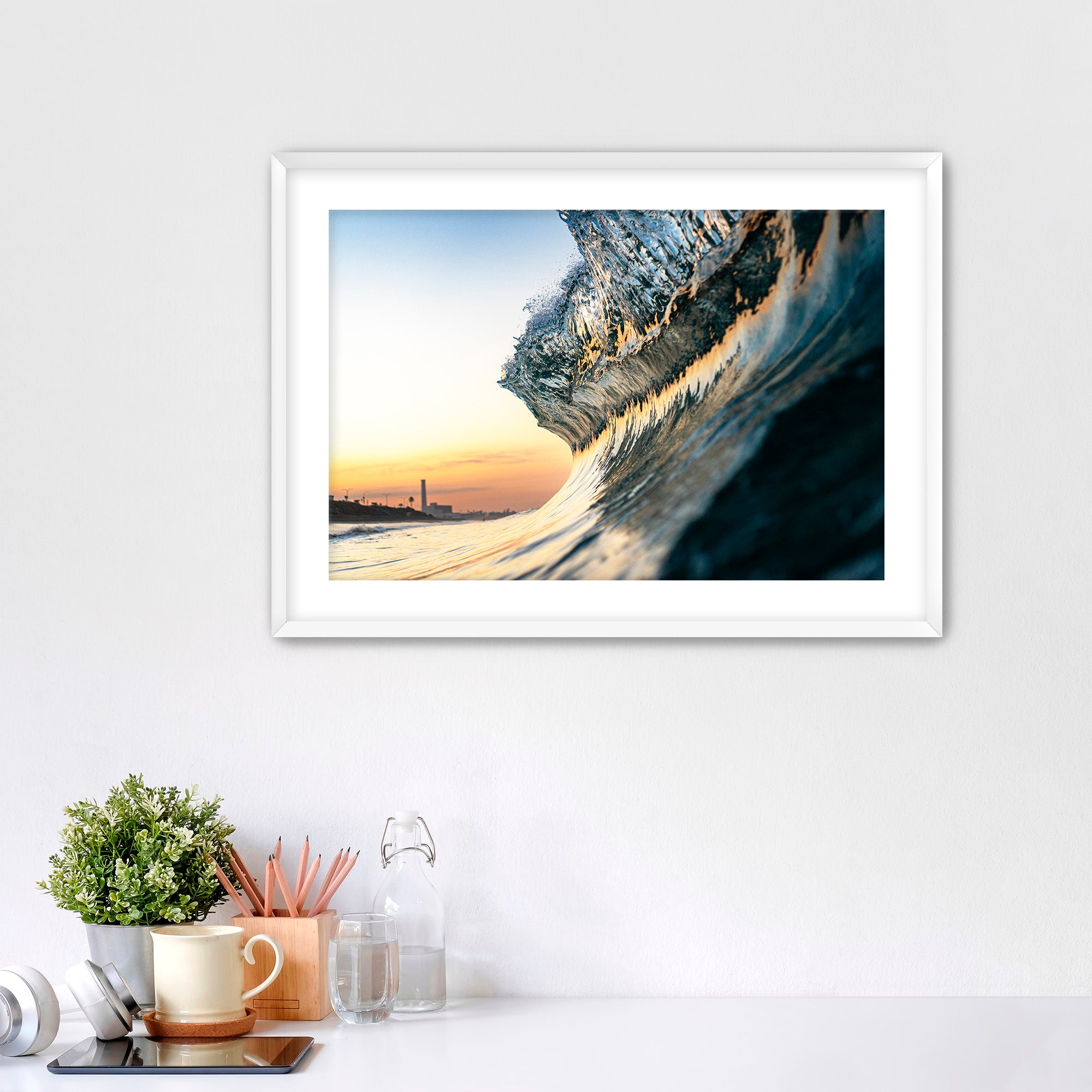 Framed photograph of a wave crashing on a beach with a power plant in the distance, displayed on a wall above a desk.