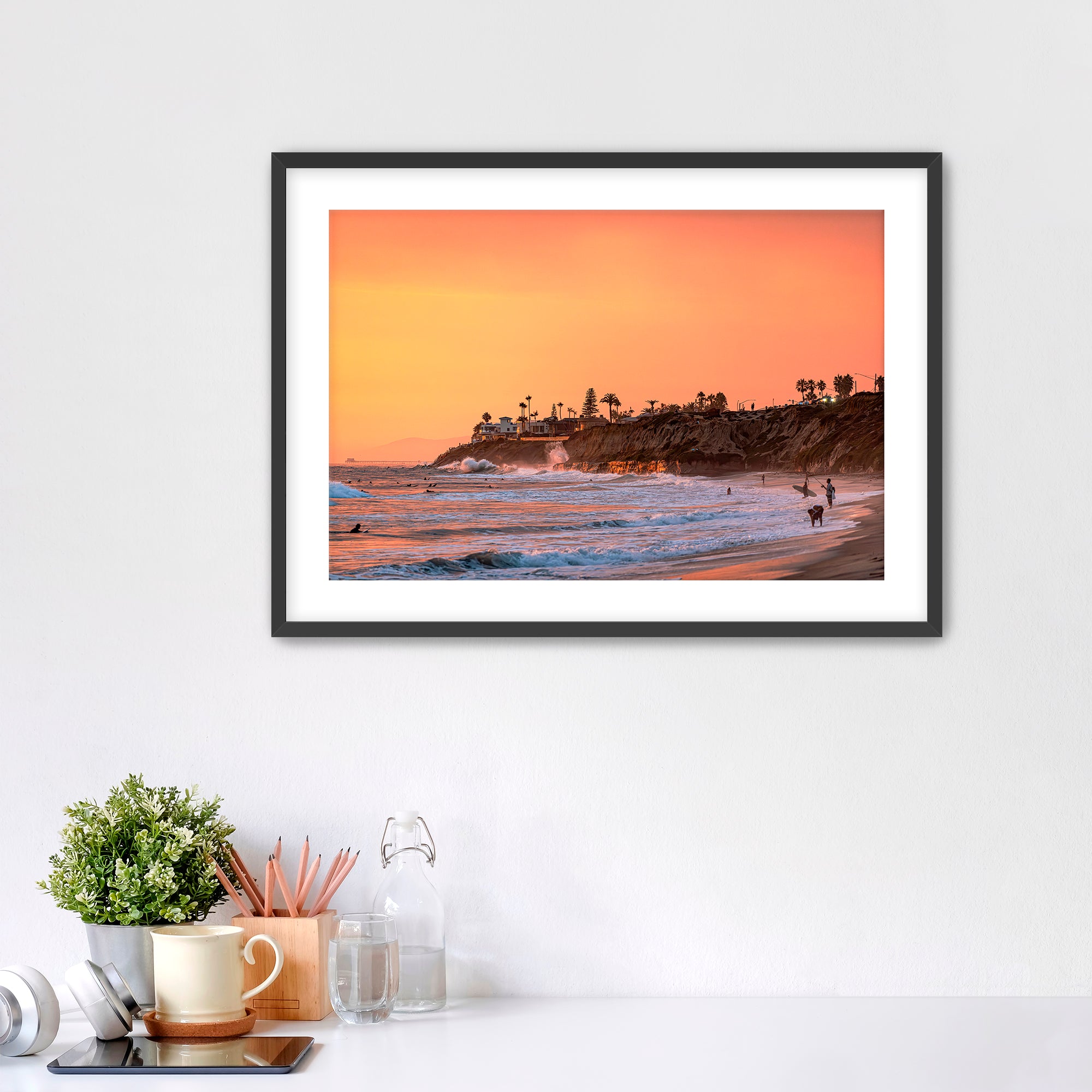 Framed artwork of a Carlsbad beach sunset on a white wall above a desk.