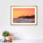 Framed artwork of a Carlsbad beach sunset on a white wall above a desk.