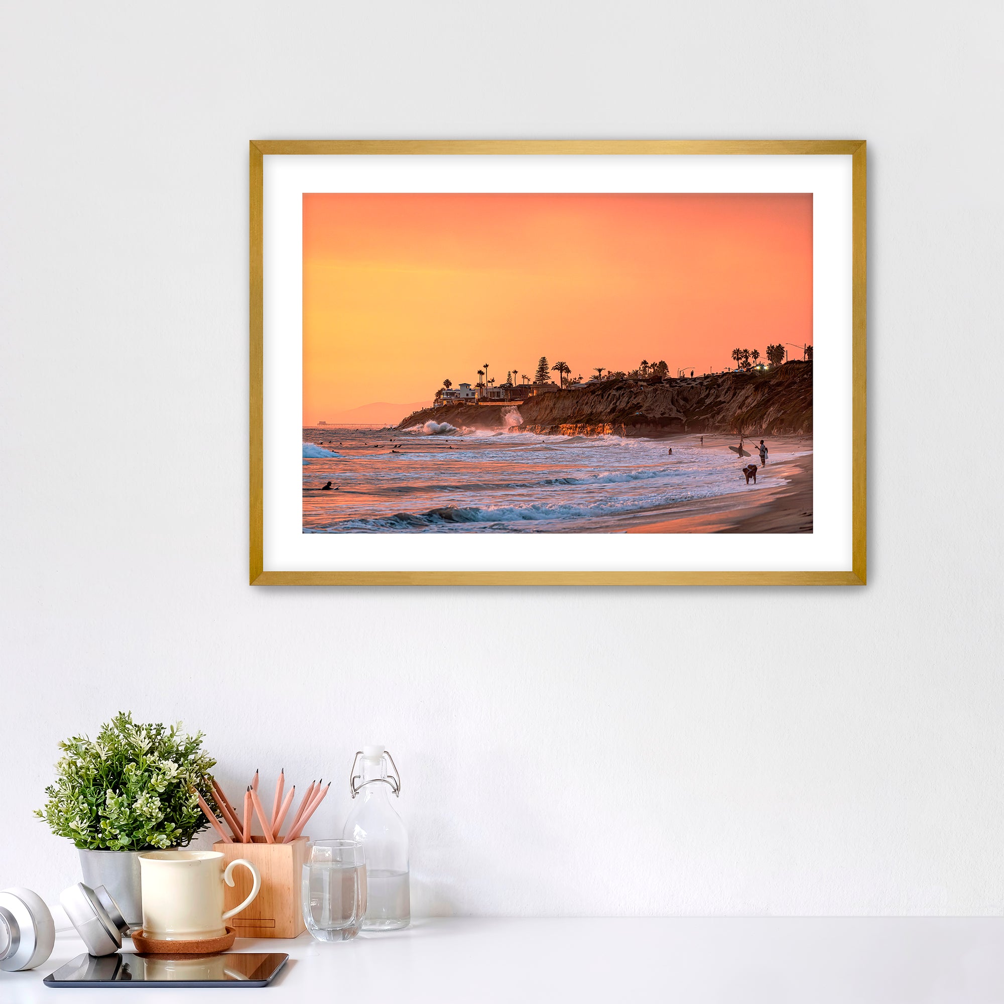 Framed artwork of a Carlsbad beach sunset on a white wall above a desk.
