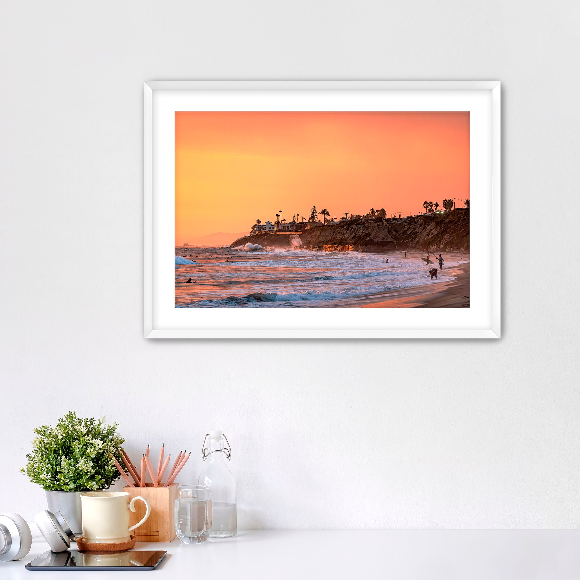 Framed artwork of a Carlsbad beach sunset on a white wall above a desk.