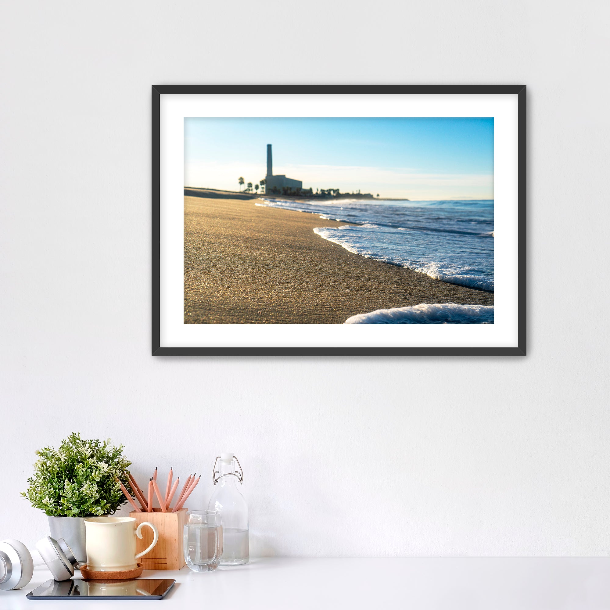 Beach scene with corduroy sand, ocean waves and a distant power plant on a white wall with desk items below. 
