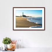Beach scene with corduroy sand, ocean waves and a distant power plant on a white wall with desk items below. 