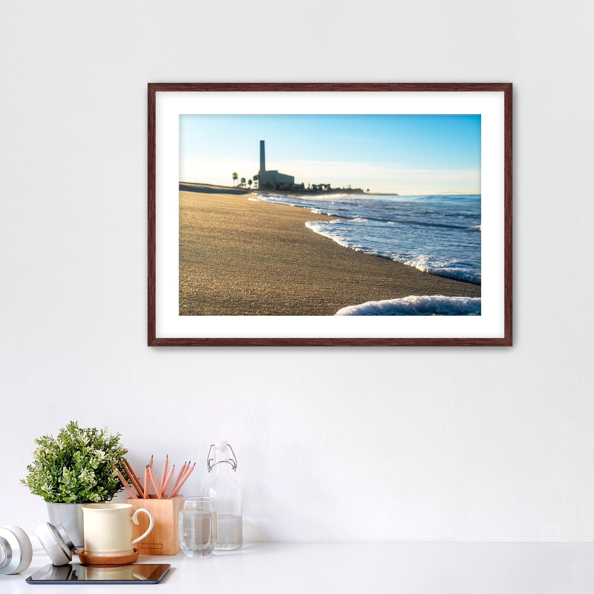Beach scene with corduroy sand, ocean waves and a distant power plant on a white wall with desk items below. 