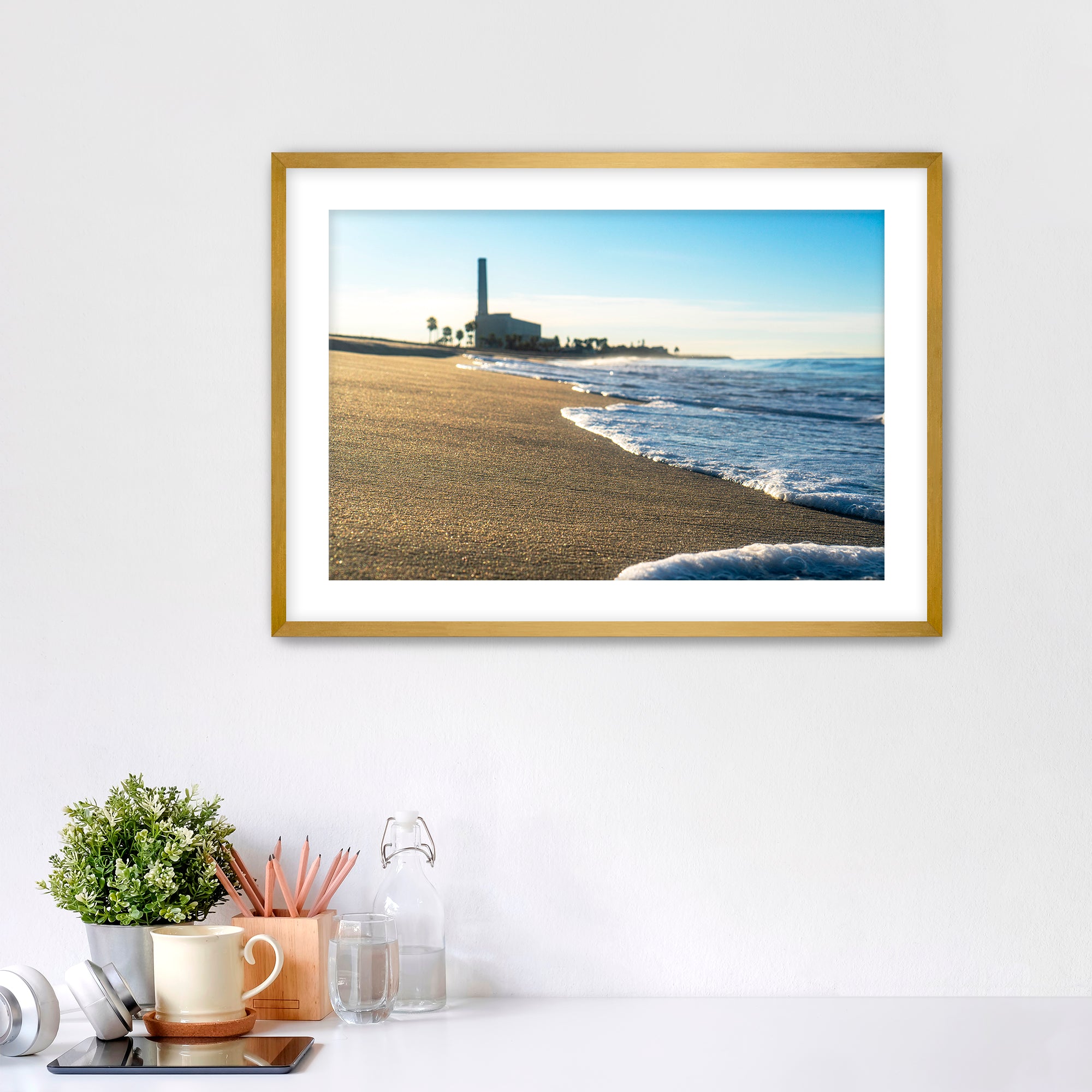 Beach scene with corduroy sand, ocean waves and a distant power plant on a white wall with desk items below. 
