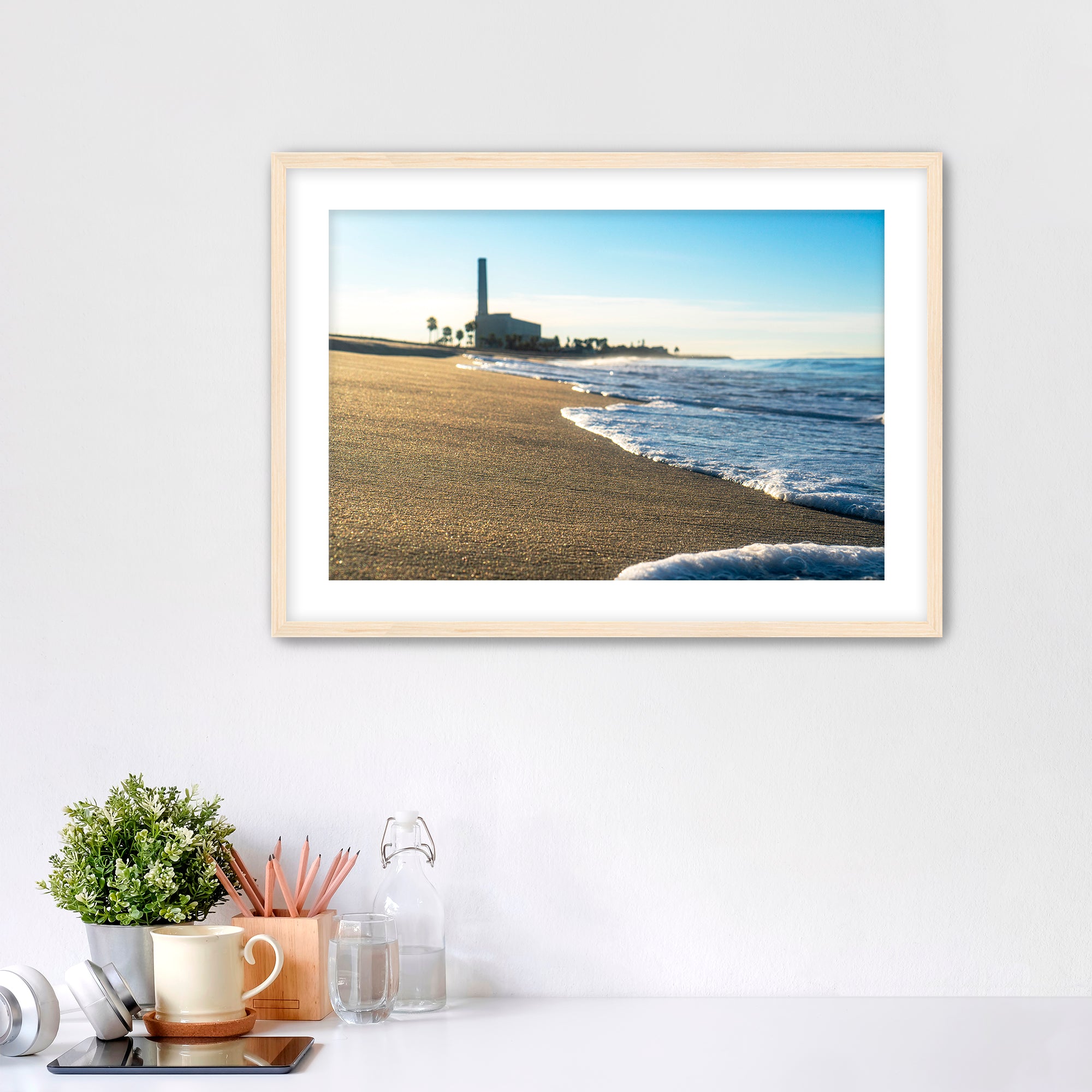 Beach scene with corduroy sand, ocean waves and a distant power plant on a white wall with desk items below. 