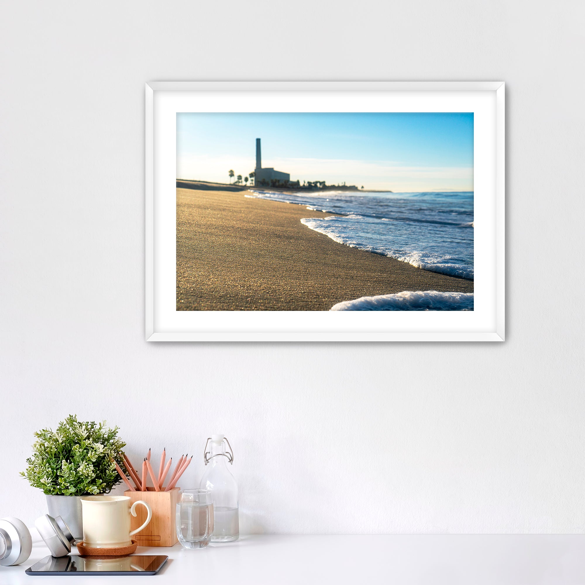 Beach scene with corduroy sand, ocean waves and a distant power plant on a white wall with desk items below. 
