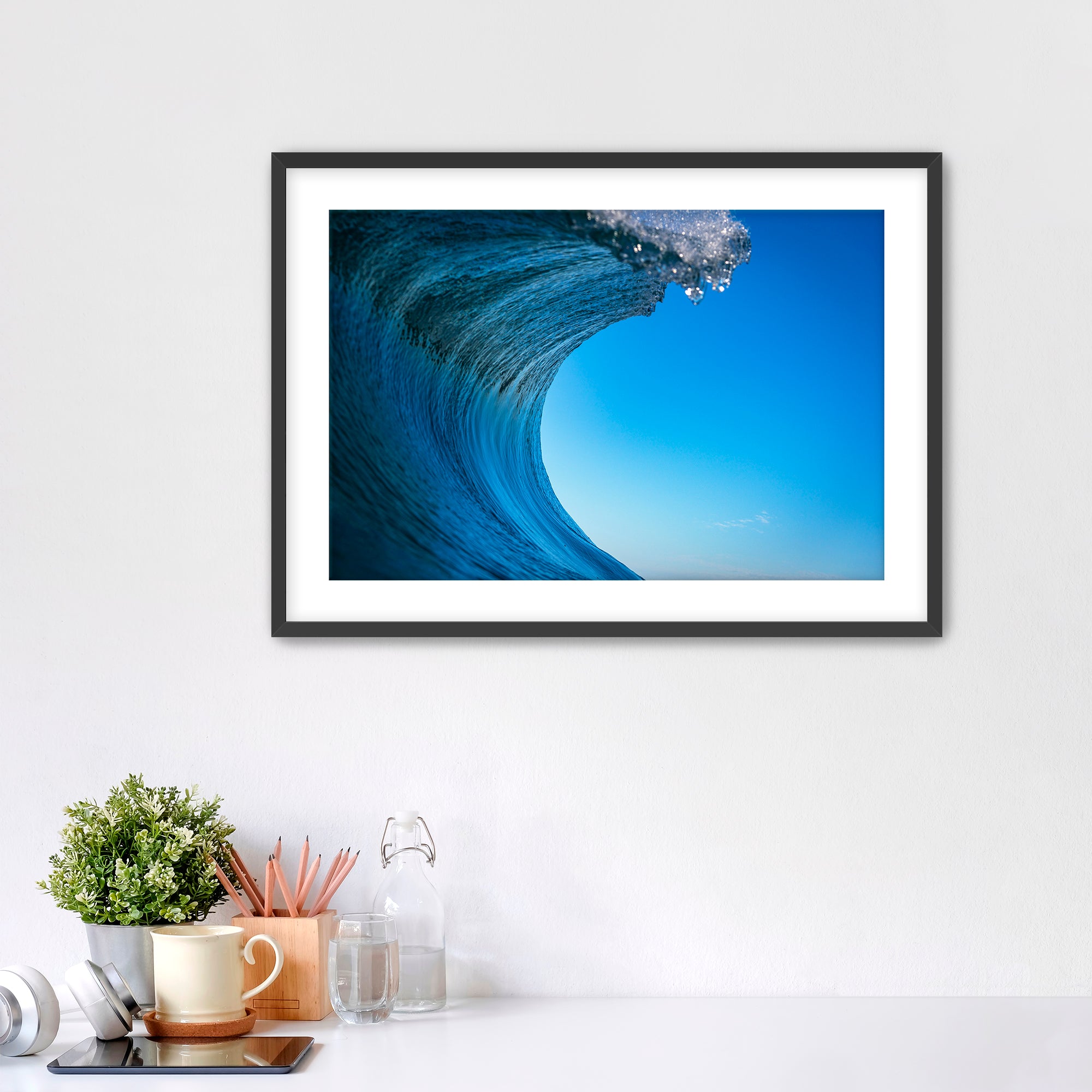 Framed print of a close-up of a curling wave with clear blue sky.  on a white wall above a desk with office supplies.