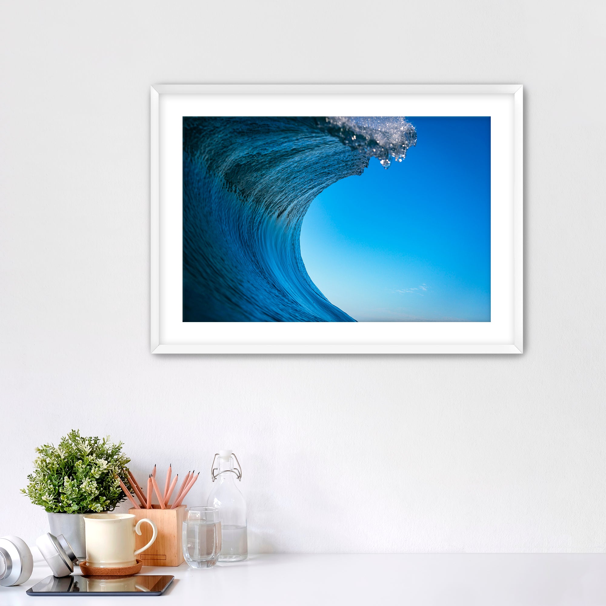 Framed print of a close-up of a curling wave with clear blue sky.  on a white wall above a desk with office supplies.