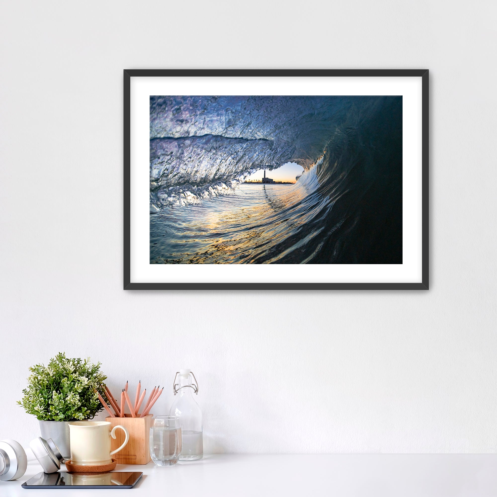 Framed artwork of a wave breaking with the Carlsbad smoke stack on a white wall above a desk with office supplies.