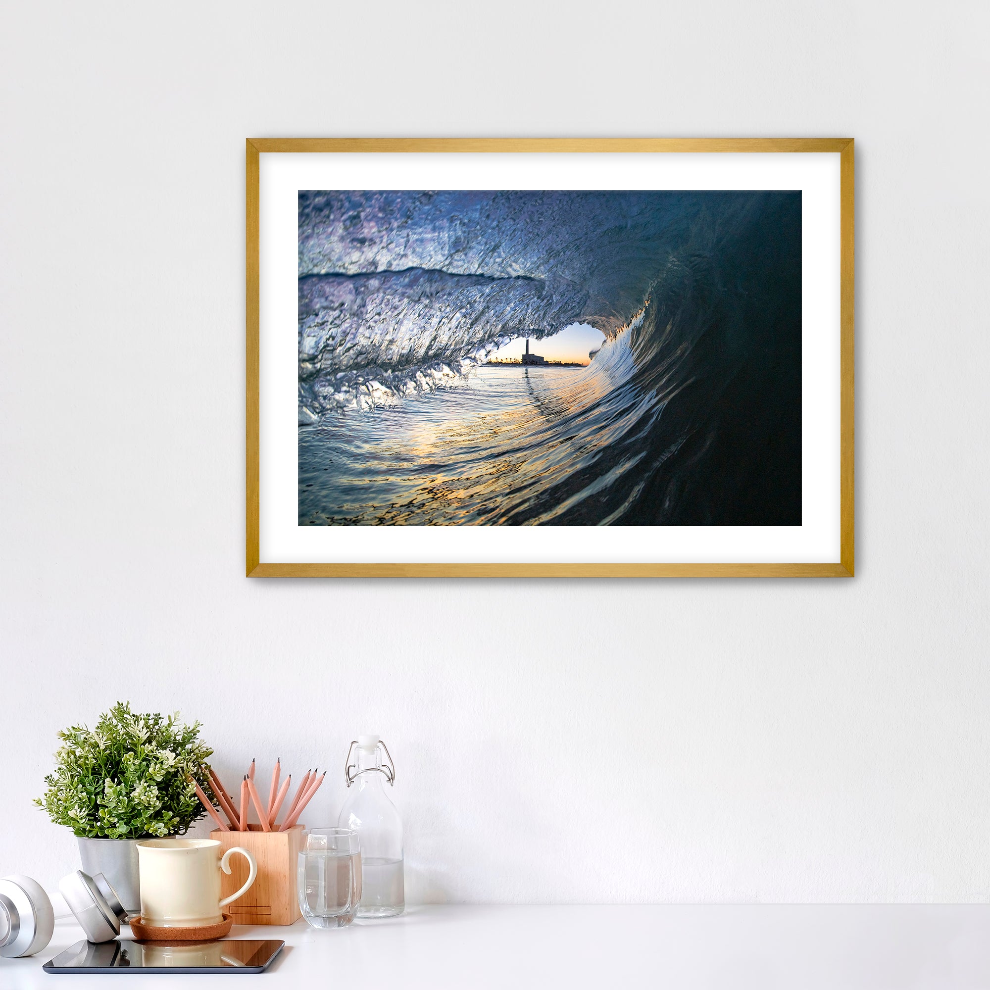 Framed artwork of a wave breaking with the Carlsbad smoke stack on a white wall above a desk with office supplies.