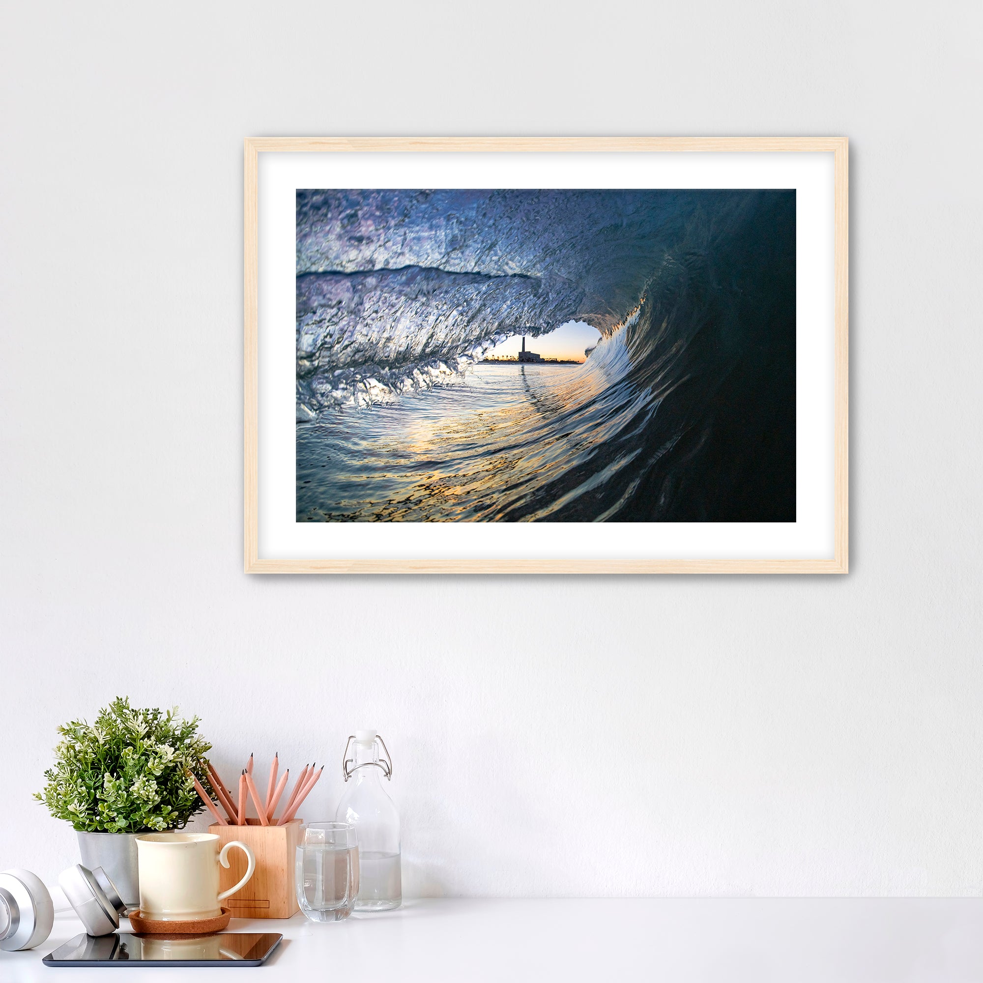 Framed artwork of a wave breaking with the Carlsbad smoke stack on a white wall above a desk with office supplies.