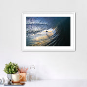 Framed artwork of a wave breaking with the Carlsbad smoke stack on a white wall above a desk with office supplies.