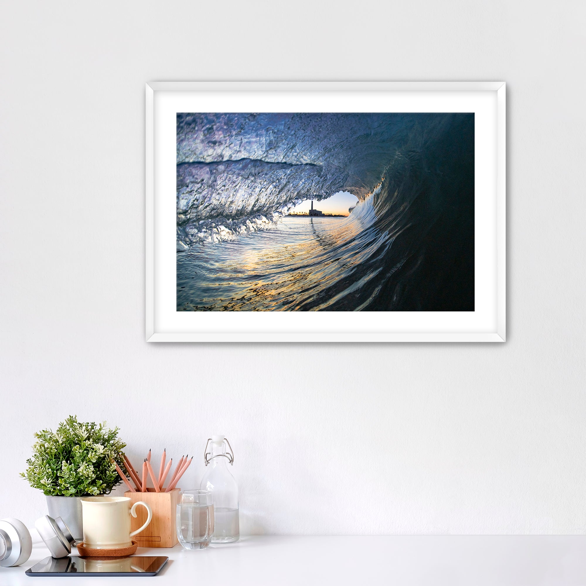 Framed artwork of a wave breaking with the Carlsbad smoke stack on a white wall above a desk with office supplies.