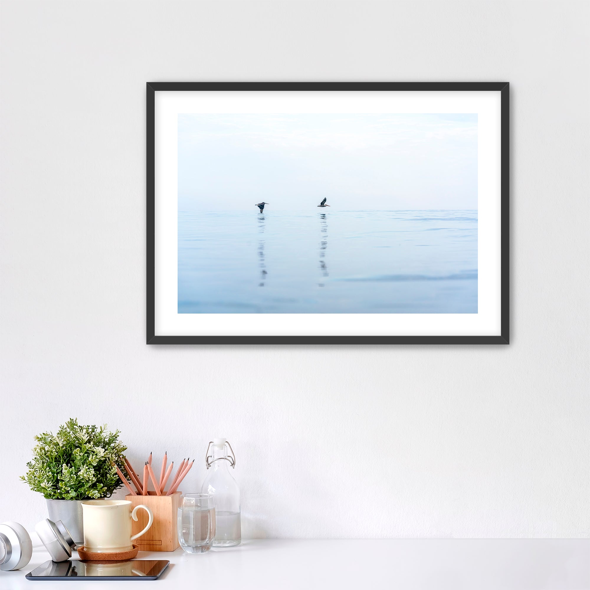 Framed artwork of two pelicans over water in Carlsbad, California, placed over a desk.