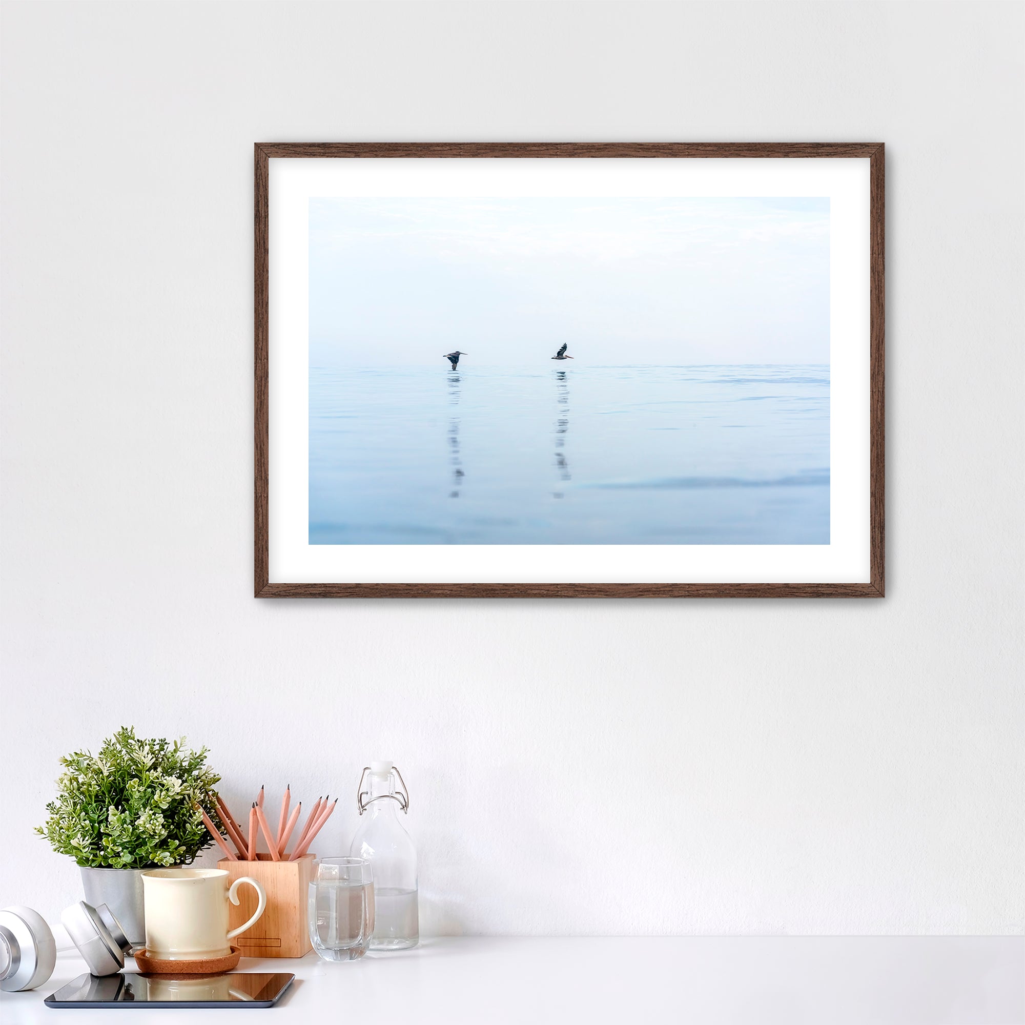 Framed artwork of two pelicans over water in Carlsbad, California, placed over a desk.