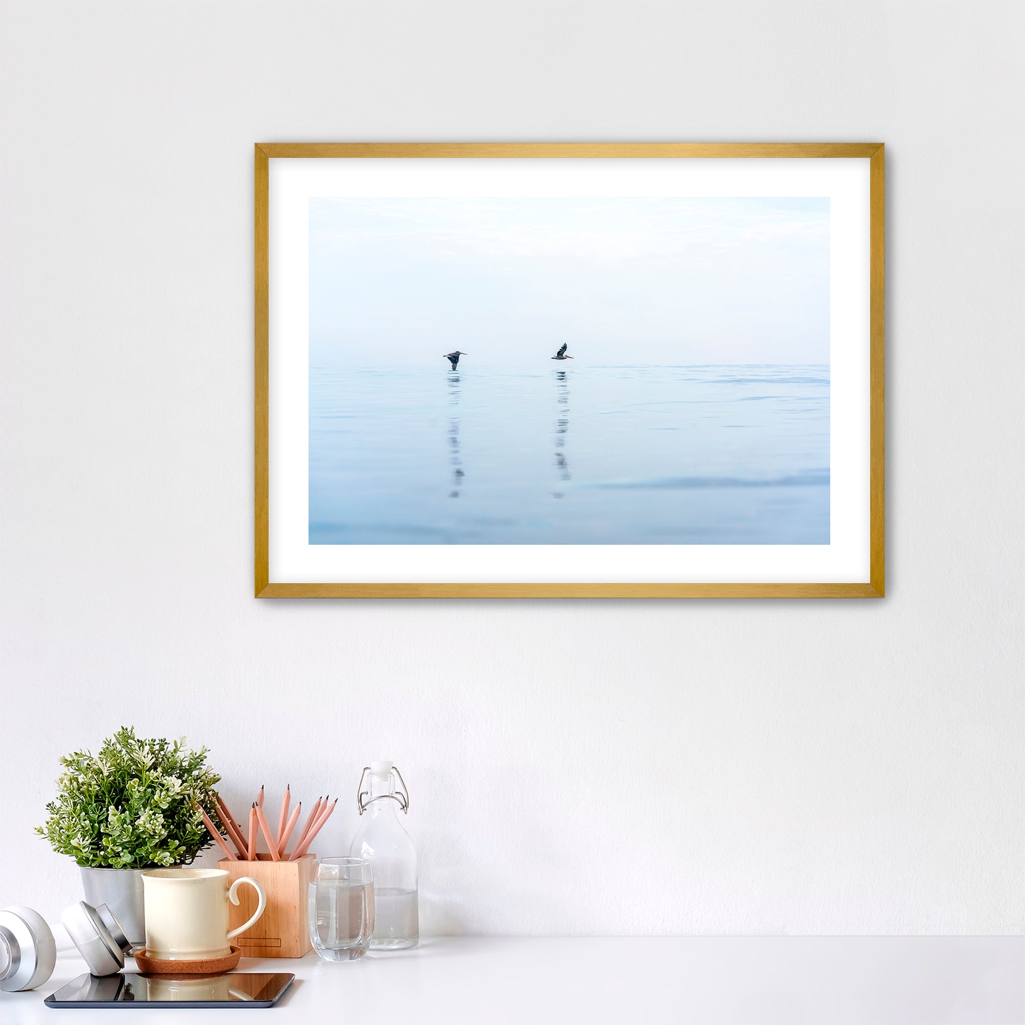 Framed artwork of two pelicans over water in Carlsbad, California, placed over a desk.