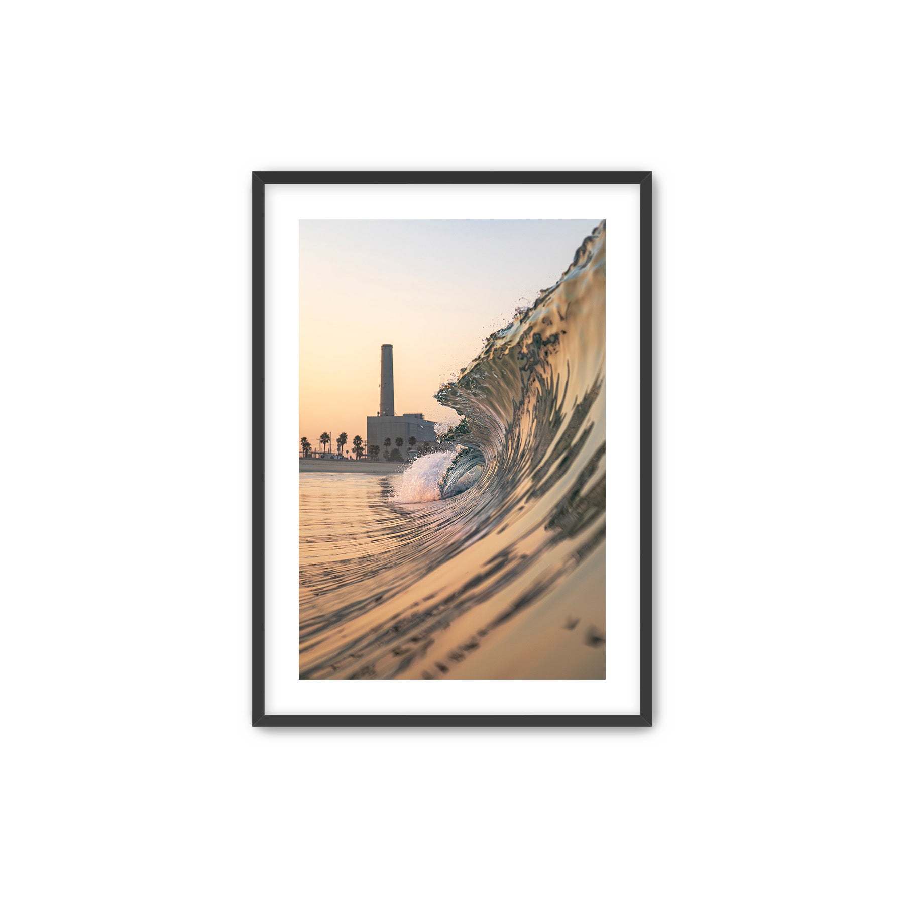Framed artwork photographic print of a wave crashing onto South Carlsbad State beach with the Carlsbad power plant in the background, displayed on a wall above a desk.