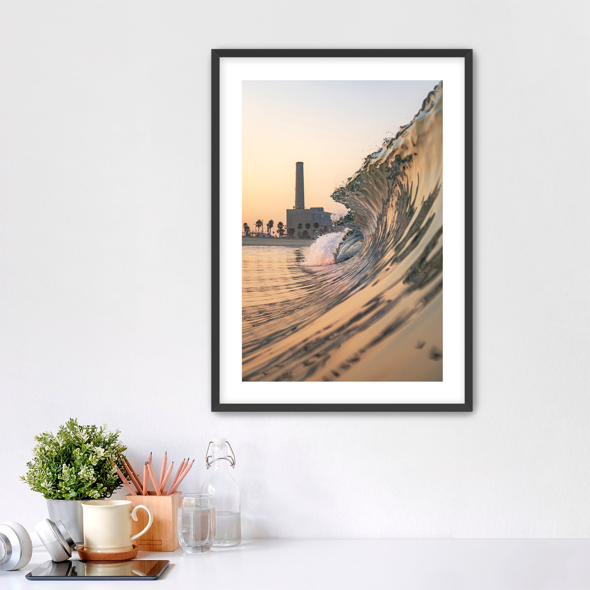 Framed artwork photographic print of a wave crashing onto South Carlsbad State beach with the Carlsbad power plant in the background, displayed on a wall above a desk.