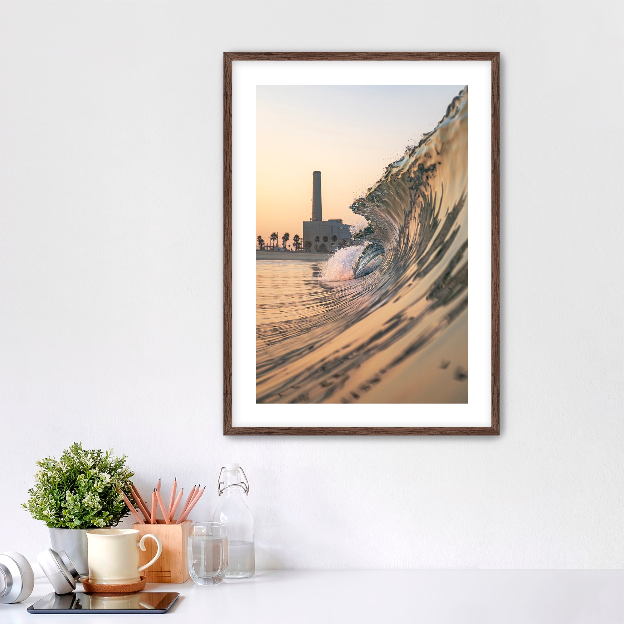 Framed artwork photographic print of a wave crashing onto South Carlsbad State beach with the Carlsbad power plant in the background, displayed on a wall above a desk.