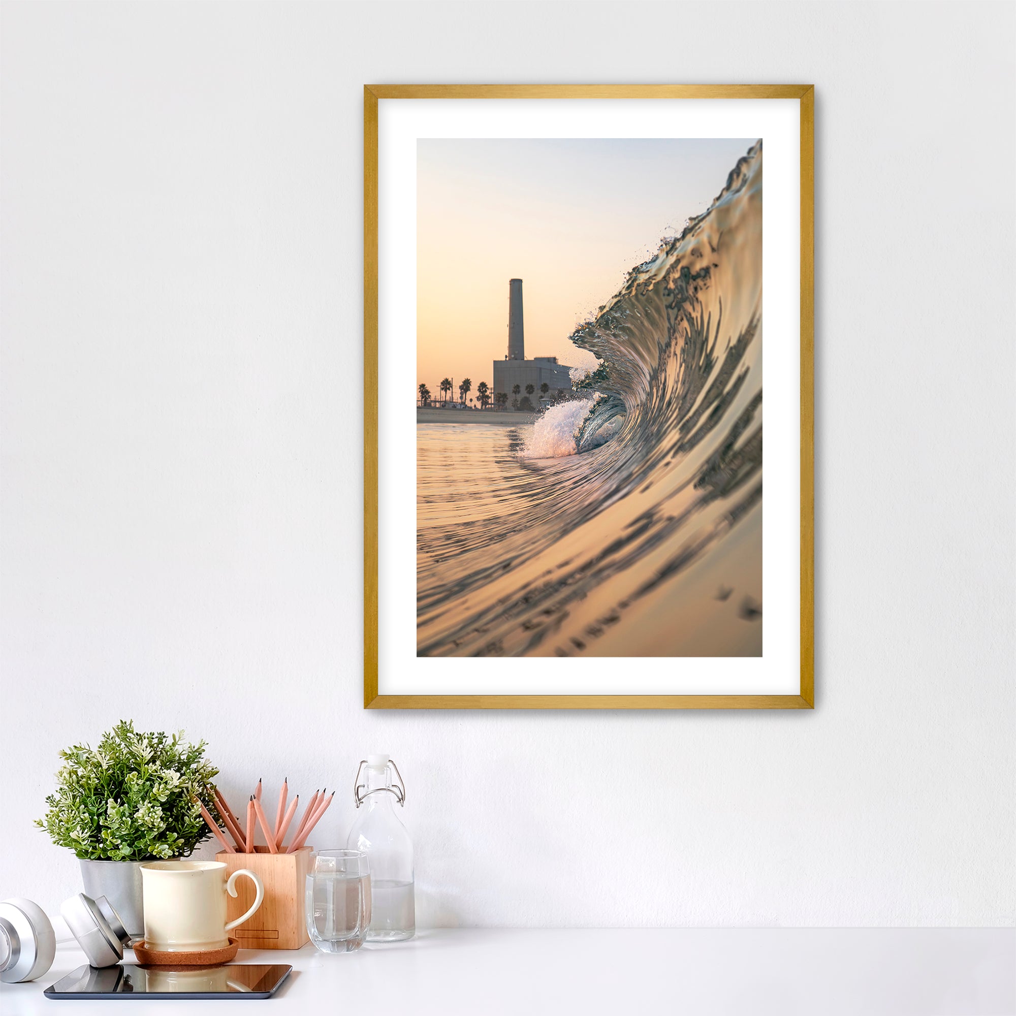 Framed artwork photographic print of a wave crashing onto South Carlsbad State beach with the Carlsbad power plant in the background, displayed on a wall above a desk.