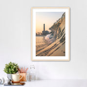 Framed artwork photographic print of a wave crashing onto South Carlsbad State beach with the Carlsbad power plant in the background, displayed on a wall above a desk.