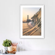 Framed artwork photographic print of a wave crashing onto South Carlsbad State beach with the Carlsbad power plant in the background, displayed on a wall above a desk.