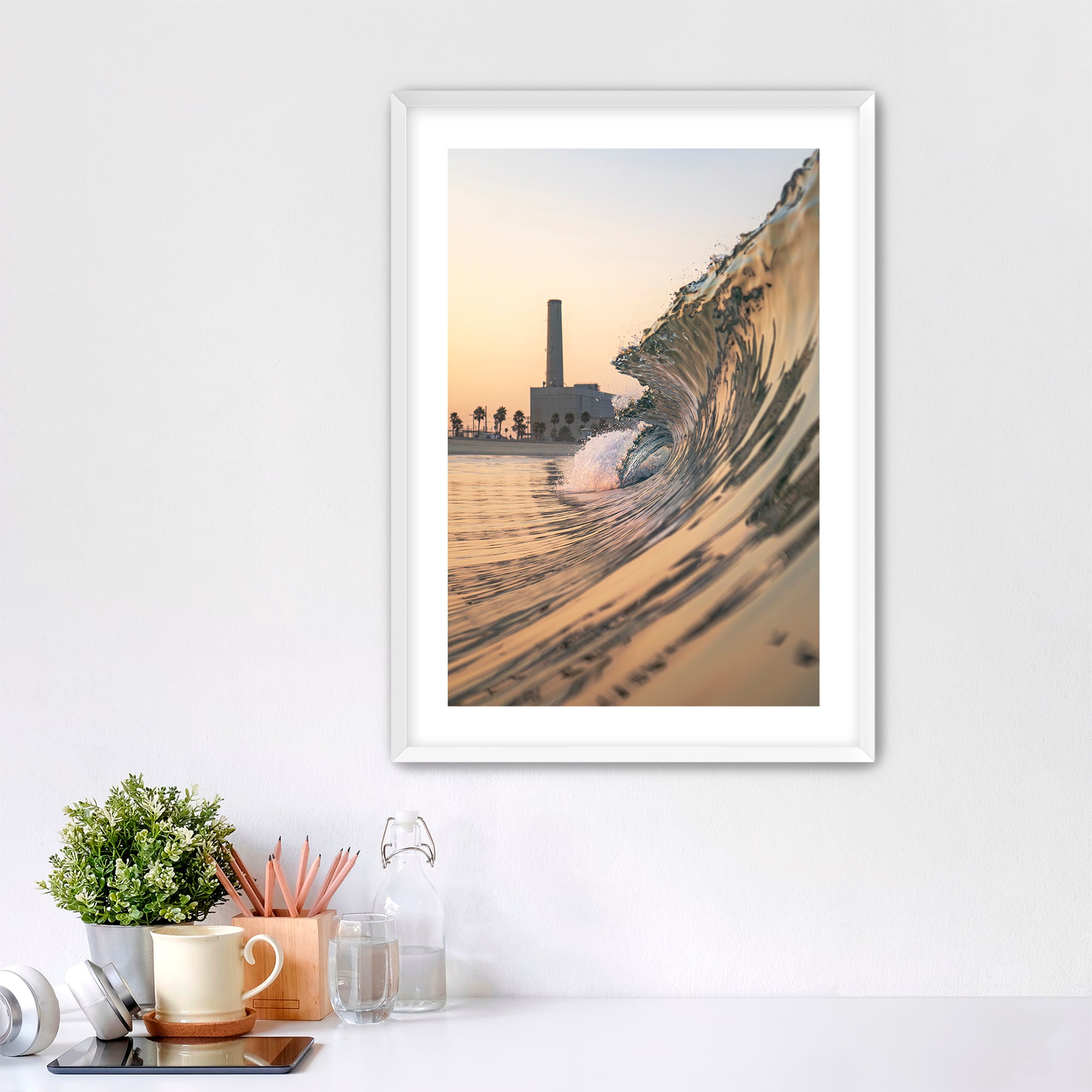 Framed artwork photographic print of a wave crashing onto South Carlsbad State beach with the Carlsbad power plant in the background, displayed on a wall above a desk.