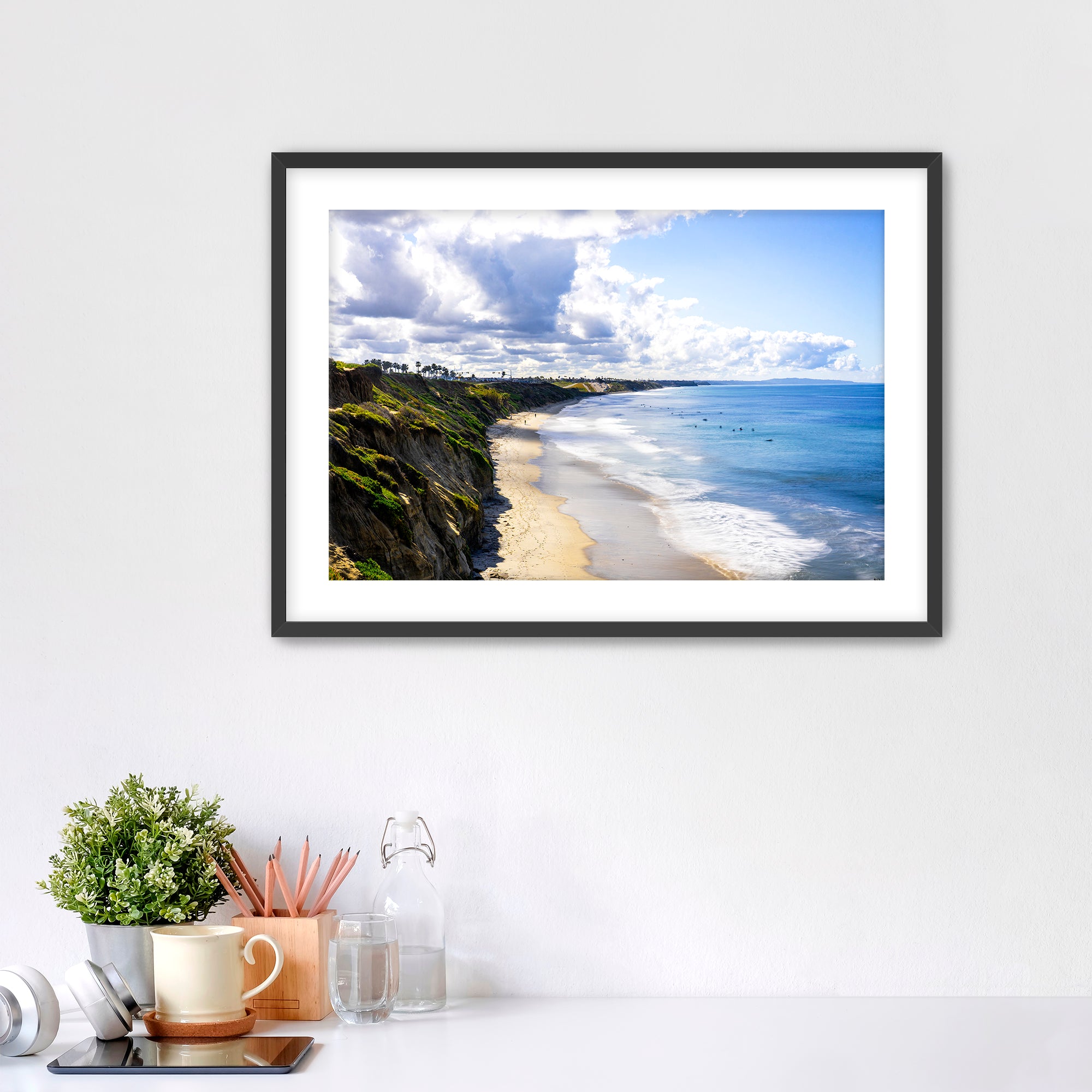 Carlsbad beach scene with cliffs and ocean under a cloudy sky on a white wall with decor items below.