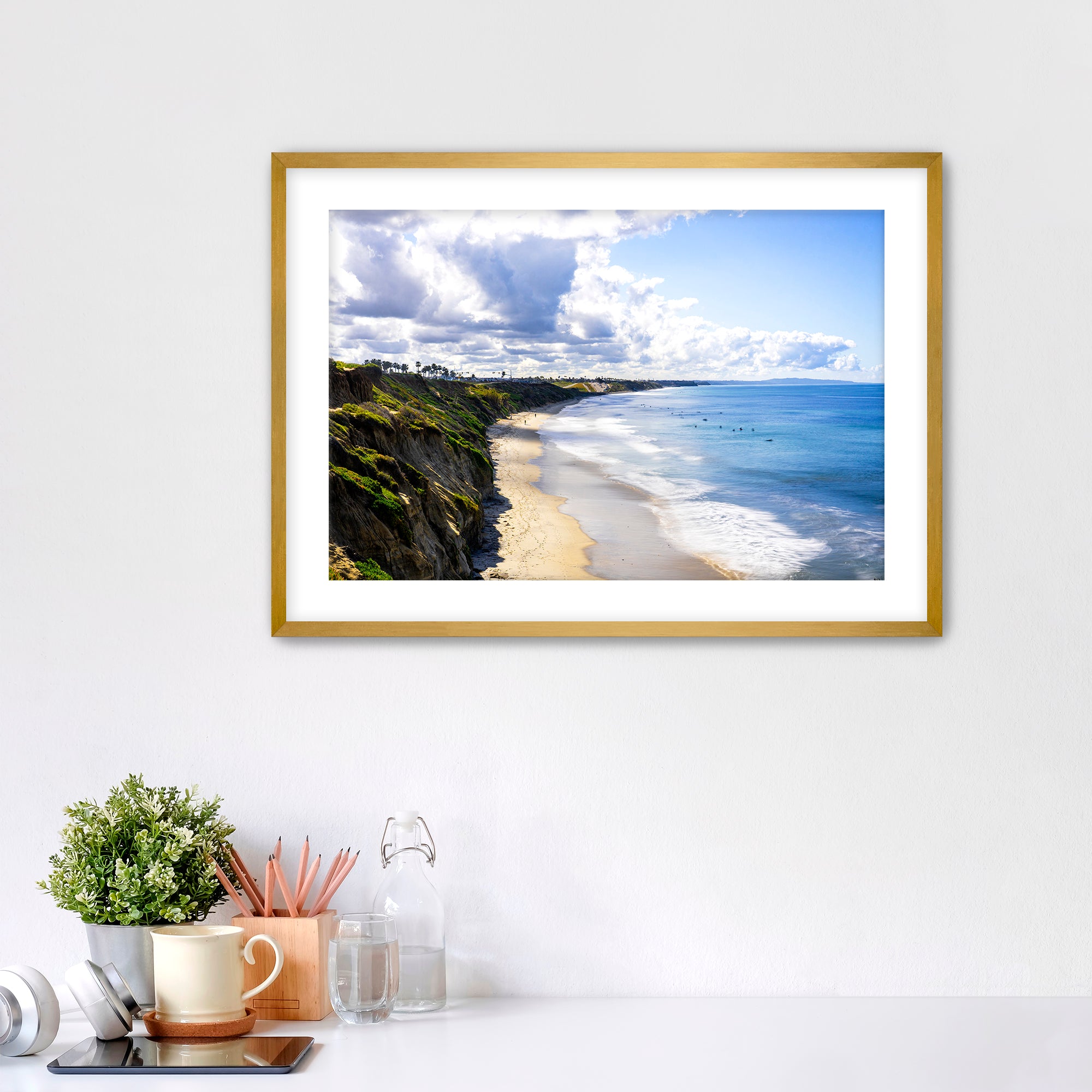 Carlsbad beach scene with cliffs and ocean under a cloudy sky on a white wall with decor items below.