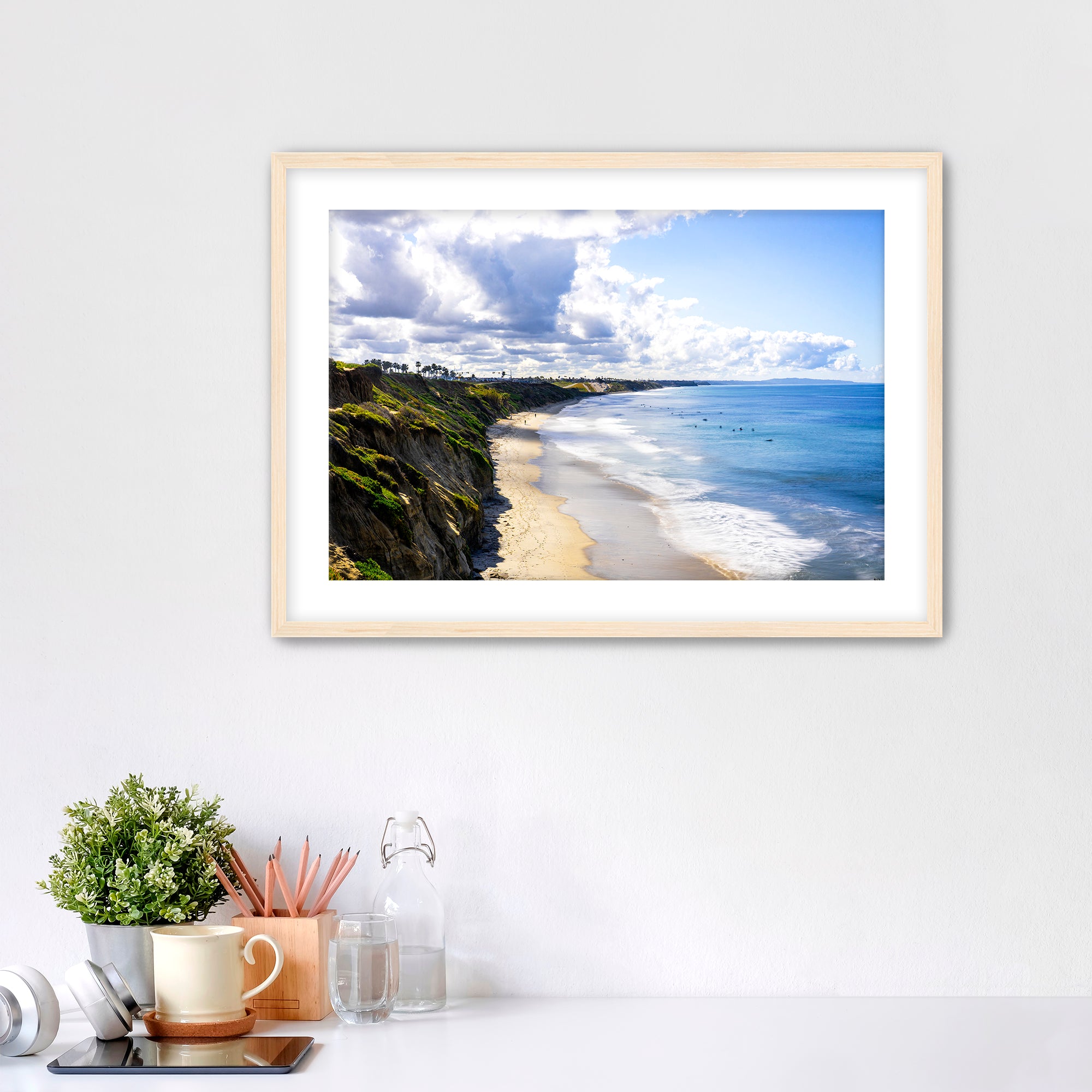 Carlsbad beach scene with cliffs and ocean under a cloudy sky on a white wall with decor items below.