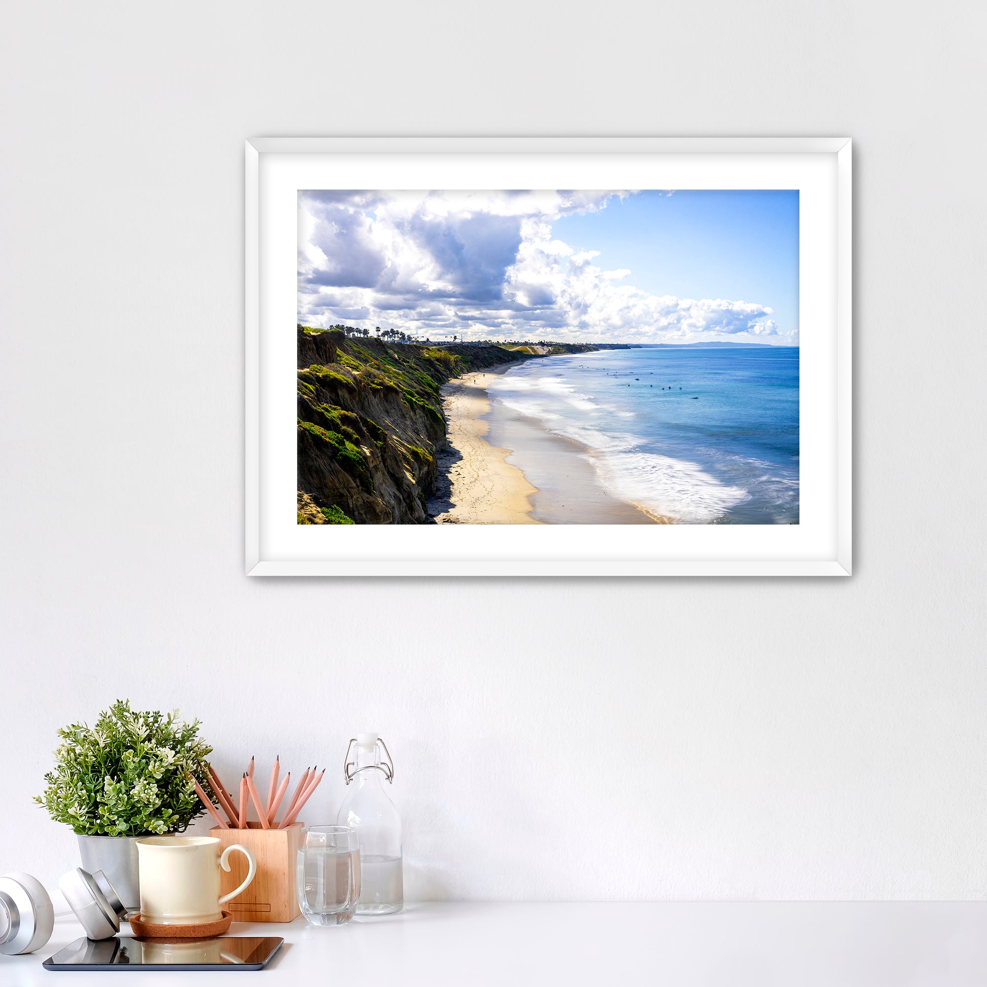 Carlsbad beach scene with cliffs and ocean under a cloudy sky on a white wall with decor items below.