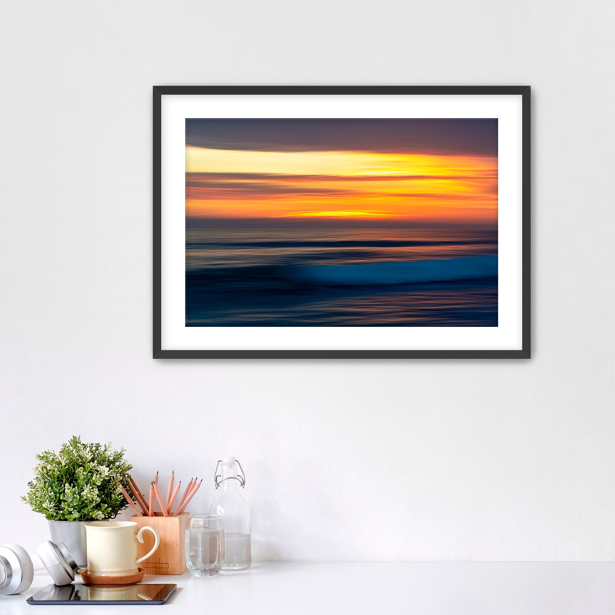 Framed artwork of a sunset over water on a white wall with decorative items below.