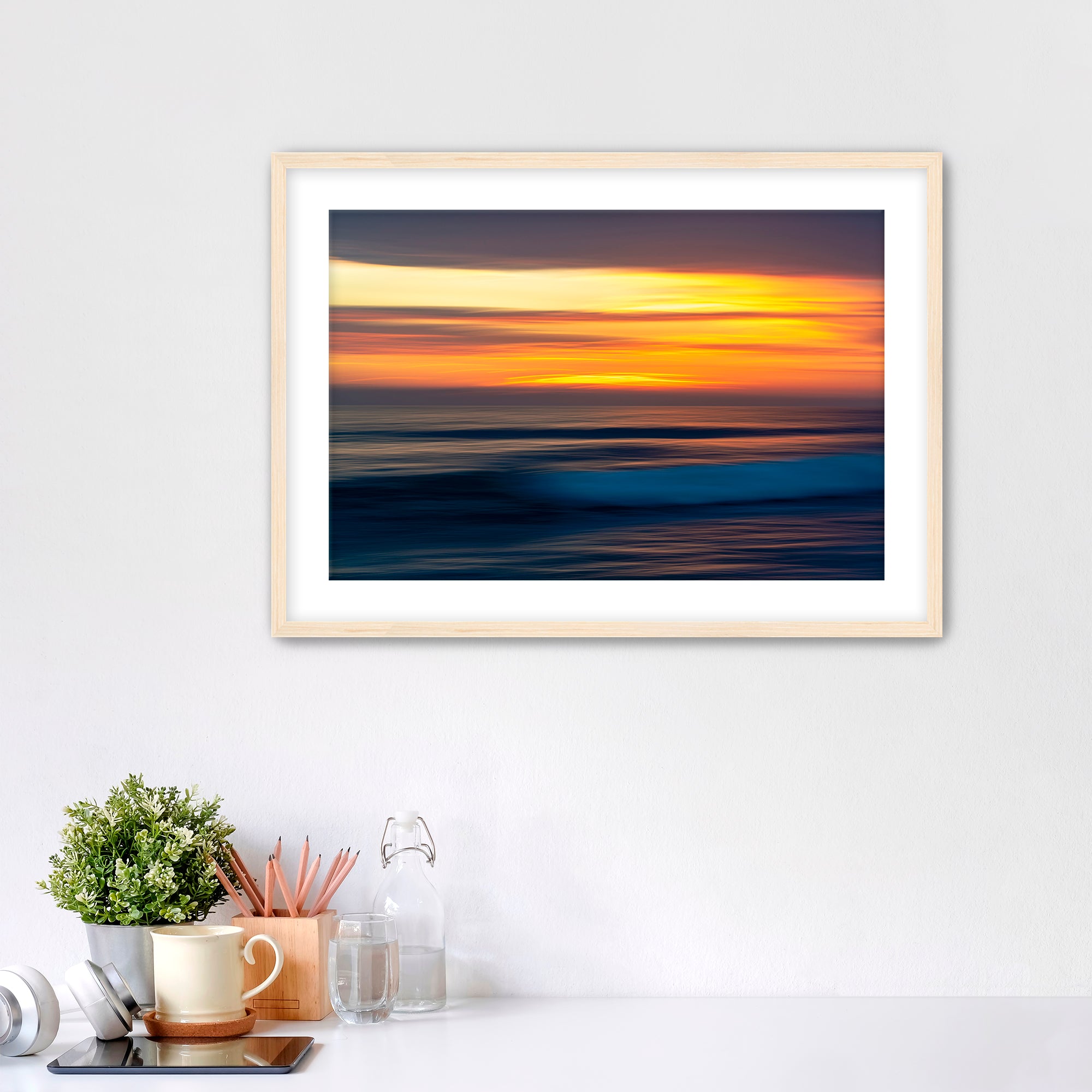 Framed artwork of a sunset over water on a white wall with decorative items below.