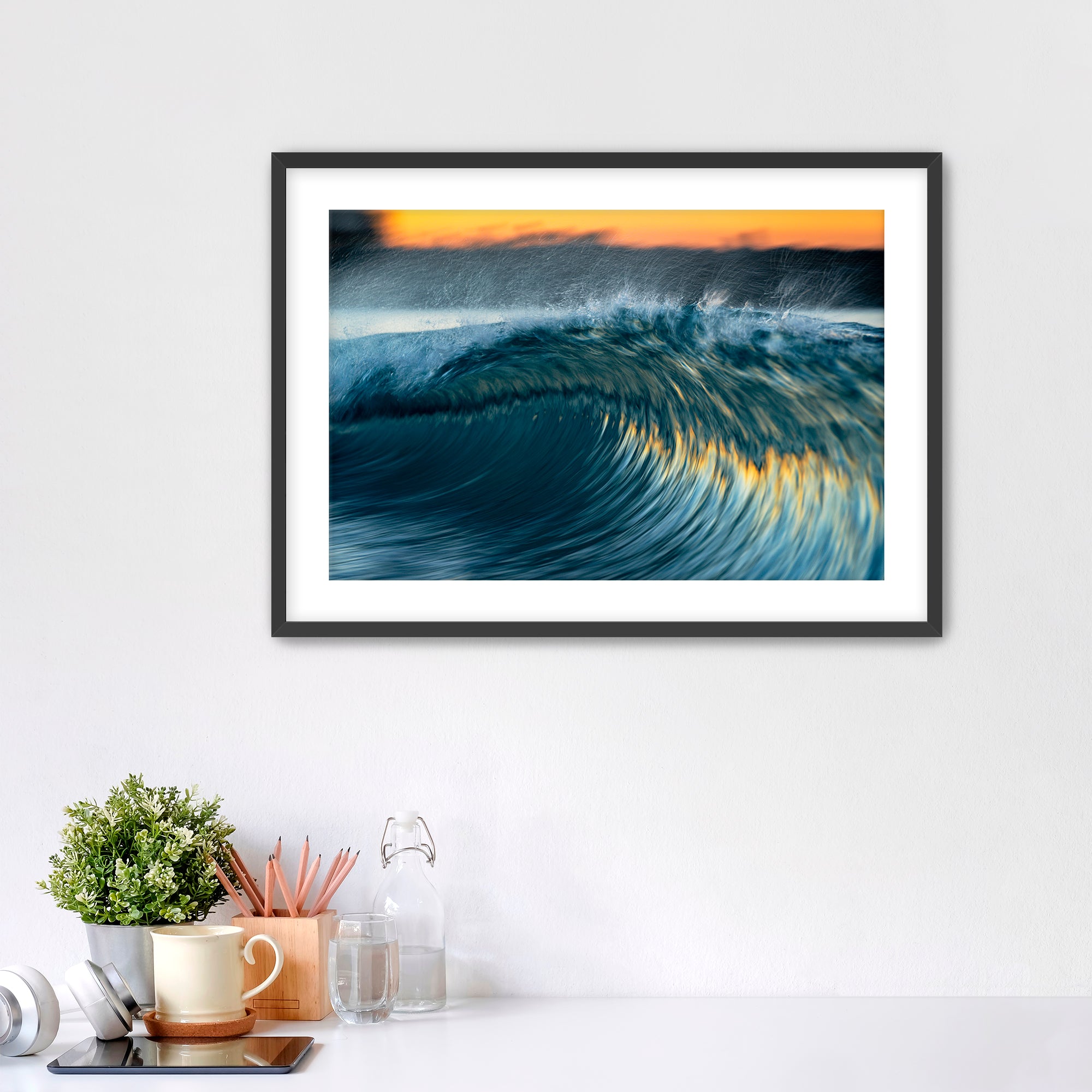 Framed photo of a colorful, slow-shutter wave on a wall above a desk with office supplies.