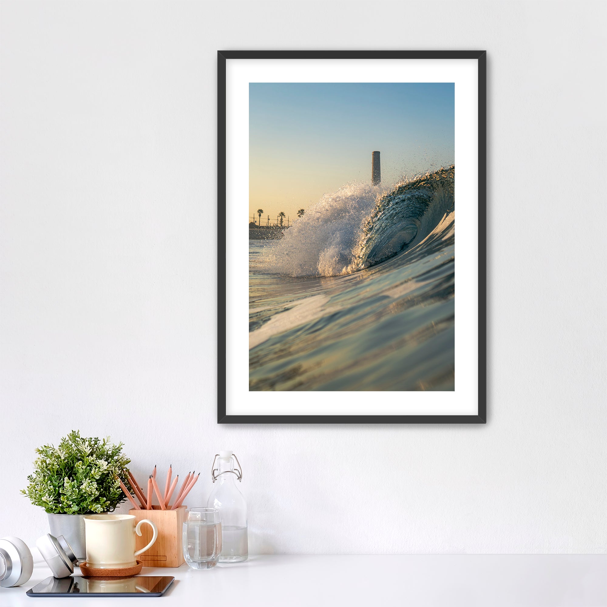 Framed photograph of a beach scene with waves crashing in front of the historic Carlsbad power plant on a white wall above a desk.