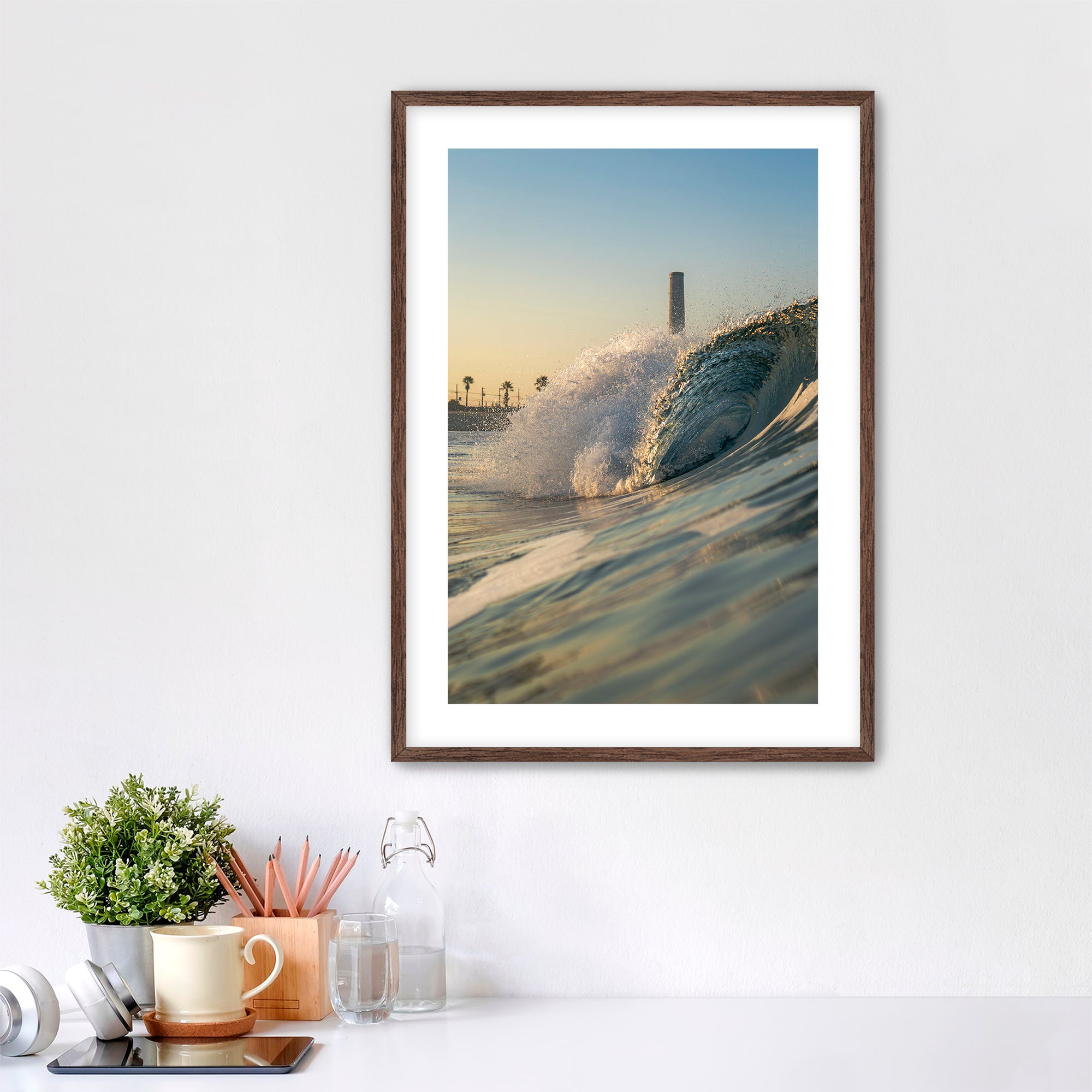 Framed photograph of a beach scene with waves crashing in front of the historic Carlsbad power plant on a white wall above a desk.
