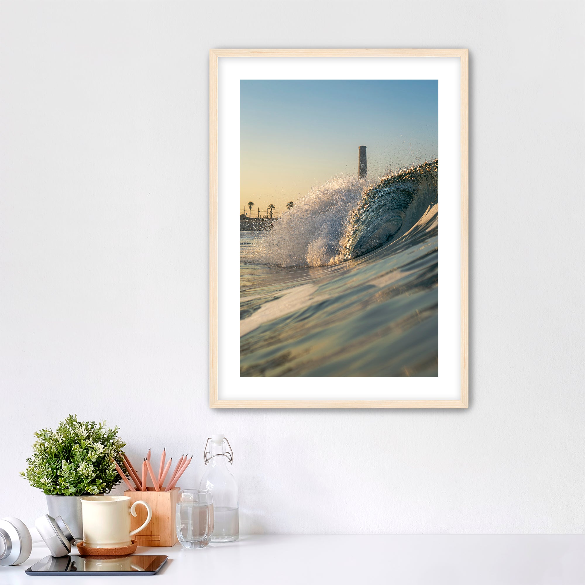 Framed photograph of a beach scene with waves crashing in front of the historic Carlsbad power plant on a white wall above a desk.