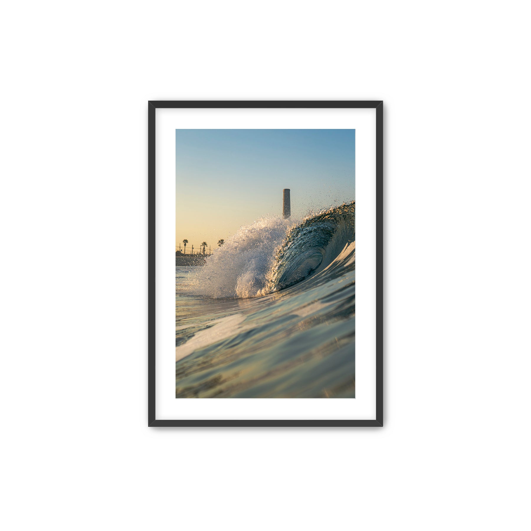 Framed photograph of a beach scene with waves crashing in front of the historic Carlsbad power plant on a white wall above a desk.