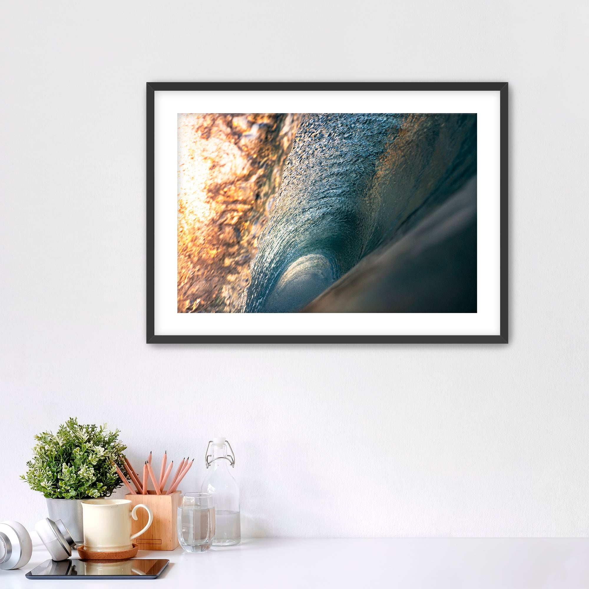 Framed photograph of the inside of a barreling wave on a white wall above a desk with office supplies.